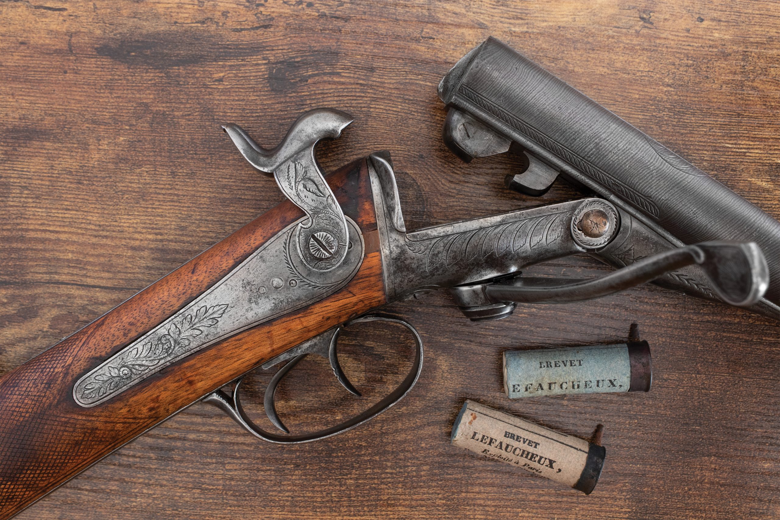 An antique Lefaucheux pinfire shotgun shown open at the breech, with two pinfire cartridges labeled “Breveté Lefaucheux” resting nearby on a wooden surface.